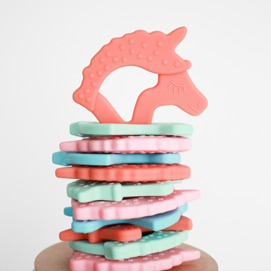 Flat stack of unicorn silicone teethers with coral unicorn upright on top