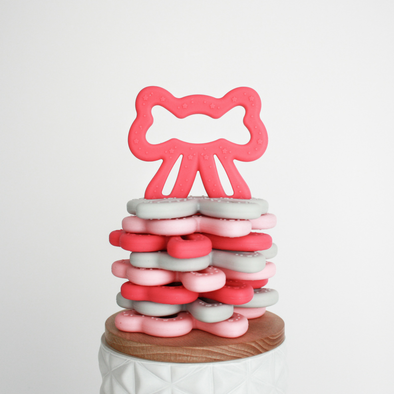 Stack of bow shaped teething toys with one coral colored bow sitting upright on top of stack
