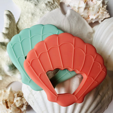Seashell teether toys. One coral, one tropical. Sand and shell background. 